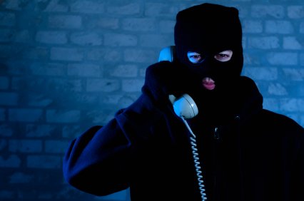 A person in all black, with a black ski mask, talking on a corded phone.