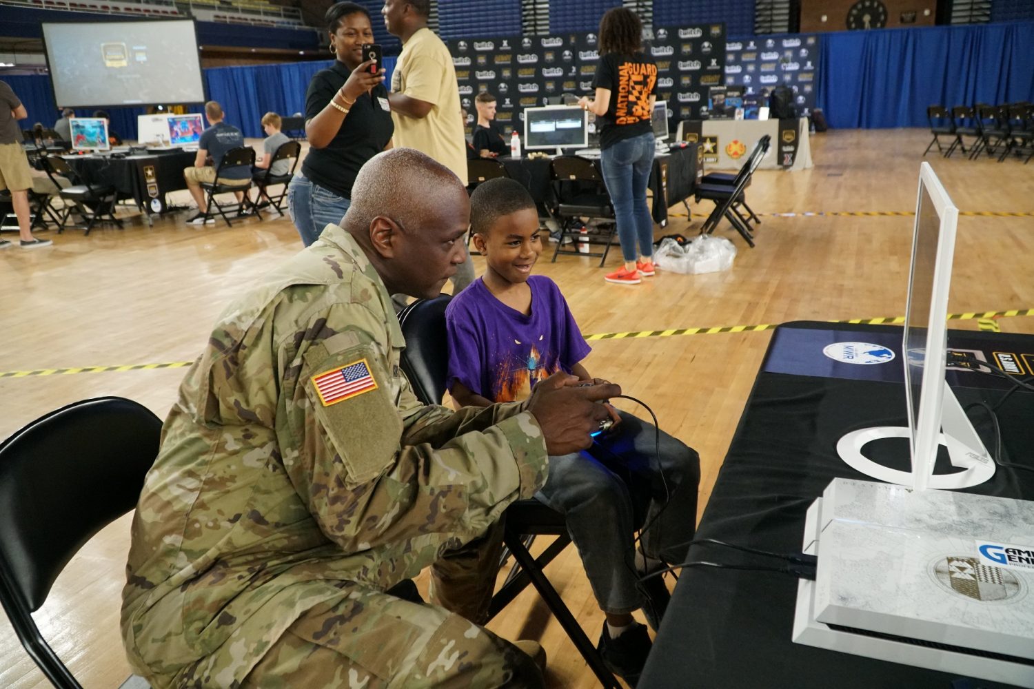 A Soldier plays a video game with a child; a woman in the background is taking a photo with her phone