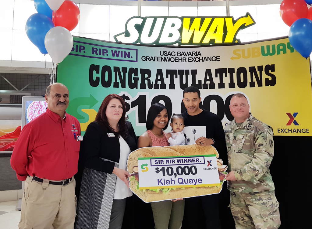 Exchange managers Aftab Khan and Carrie Cammel, along with USAG Bavaria CSM Micheal Sutterfield USAG Bavaria Command Sergeant Major present the $10,000 gift card to Kiah Quay and husband SPC Damion Quaye.