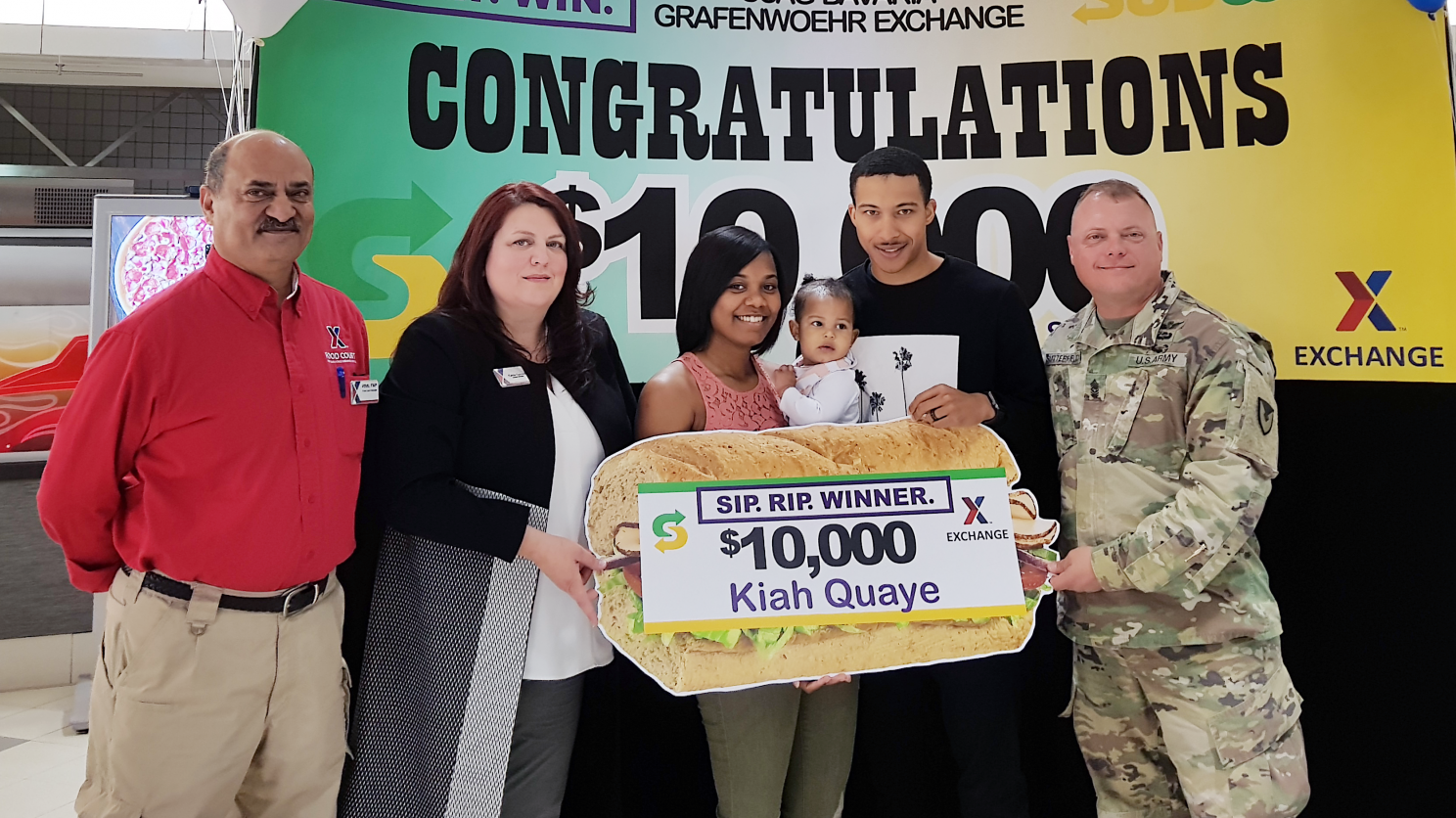 Exchange managers Aftab Khan and Carrie Cammel, along with USAG Bavaria CSM Micheal Sutterfield USAG Bavaria Command Sergeant Major present the $10,000 gift card to Kiah Quay and husband SPC Damion Quaye.