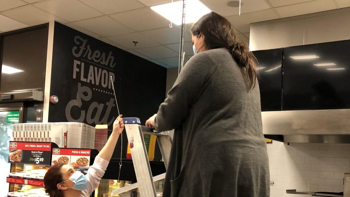 From left, Vicenza Exchange visual merchandisers Keti Nani and Kimberlyn Lastimosa set up a protective shield to hang between customers and cashiers at Hunt Brothers Pizza. They also developed a social distancing campaign to include floor signs that remind customers to stay at least one meter (6 feet) apart while shopping—a campaign that has since been adopted at Exchanges worldwide.