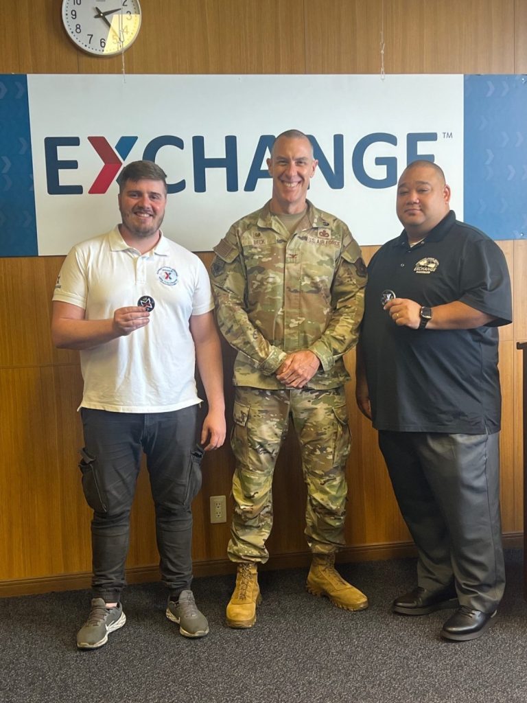 Repair & Maintenance Supervisor Dimitrios Diamantopoulos, Pacific Region Commander Col. Jason Beck and Food Plant Operations Manager Uji Battle in front of Exchange sign. 